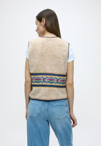 Μπεζ γιλέκο από φλις με πολύχρωμο γεωμετρικό σχέδιο στη μέση και μπλε λεπτομέρειες. Φορεμένο πάνω από λευκό μπλουζάκι με ανοιχτό μπλε τζιν.