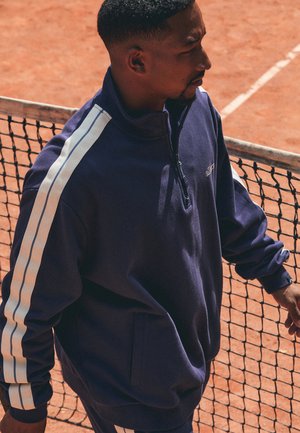 Navy blue, zip-up sweatshirt with white side stripes and a stand-up collar. Made from soft material with a relaxed fit and front pockets.