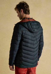 Mężczyzna w ciemnoniebieskiej pikowanej kurtce z kapturem, nałożonej na brązową koszulę w kratę, w połączeniu z czerwonymi spodniami, stojący na jednolitym beżowym tle.