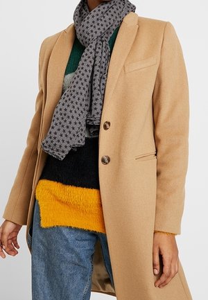 Persoon draagt een beige jas, een grijze sjaal met sterrenpatroon, een trui in kleurvlakken van groen, zwart en geel, en een blauwe spijkerbroek tegen een effen achtergrond.