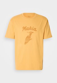 Gul bomull T-skjorte med et hakkespett-grafikk og "Makia" tekst i en kontrasterende farge. Korte ermer og rund hals.