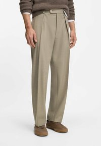 Beige geplooide broek met een getailleerde pasvorm. Gemaakt van textuurstof, met een knoopsluiting en zijzakken. Gepaard met bruine sneakers.