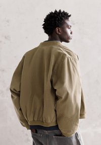 Giacca bomber verde oliva con orlo e polsini a coste, caratterizzata da una vestibilità rilassata e una texture liscia, indossata sopra una camicia blu navy.