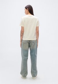 Μπεζ βαμβακερό t-shirt με λαιμόκοψη, συνδυασμένο με ανοιχτό μπλε, φαρδιά τζιν παντελόνια. Η πίσω όψη αποκαλύπτει έναν απλό σχεδιασμό χωρίς ιδιαίτερα χαρακτηριστικά.