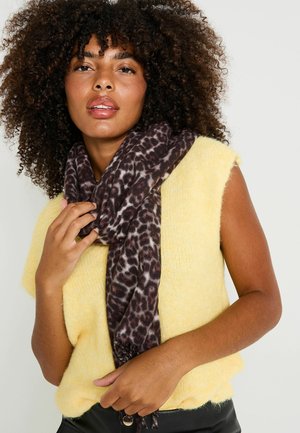 Bufanda de estampado de leopardo en marrón oscuro y crema, anudada alrededor del cuello, combinada con un suéter sin mangas de color amarillo suave.
