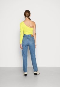 Kvinna vänd bort med ryggen i en neon-gul one-shoulder topp, blå högmidjade jeans, vita klackstövlar och hoopsringar.