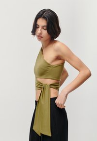 Olive green, one-shoulder top with a knot detail at the waist. Made from a smooth, stretchy material. Paired with black wide-leg pants.