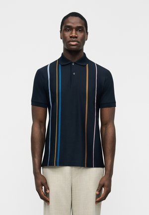 Άνδρας που φοράει κοντομάνικο μπλε-σκουρο πόλο μπλουζάκι με κάθετες μπλε, καφέ και ροζ ρίγες, συνοδευόμενο από ανοιχτό μπεζ παντελόνι.