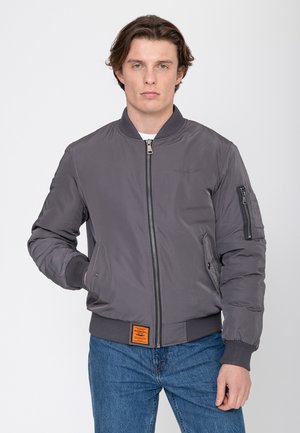 Junger Mann trägt eine graue Bomberjacke mit gerippten Bündchen und Kragen, Hände in den Taschen, kombiniert mit blauen Jeans vor einfachem Hintergrund.