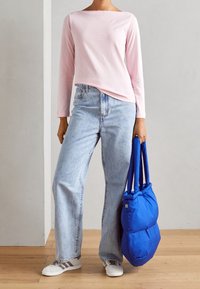Helles pinkes Langarmhemd, hochgeschnittene hellblaue Jeans und weiße Schuhe mit grauen Akzenten. Hält eine blaue gepolsterte Tasche mit zwei Griffen.