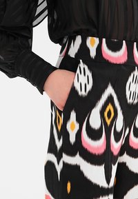 Main dans la poche d'un pantalon noir orné de motifs abstraits audacieux en blanc, rose et jaune, associée à une manche de blouse noire transparente à rayures.