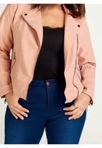 Różowa kurtka ze skóry ekologicznej z zamkiem na przodzie i żebrowanymi detalami na ramionach, noszona na czarnej bluzce i zestawiona z ciemnoniebieskimi dżinsami.