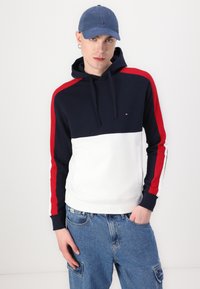 Hooded sweatshirt in navy, white, and red with a color-block design. Features a front kangaroo pocket and ribbed cuffs. Paired with denim shorts.