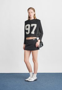 Μαύρο δίχτυ crop top με λευκό νούμερο 97, συνδυασμένο με μια μαύρη μίνι φούστα. Τα αξεσουάρ περιλαμβάνουν μια μικρή μαύρη τσάντα και ασημένια υποδήματα με λευκές κάλτσες.