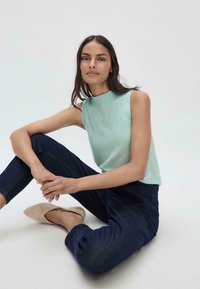 Lichtblauw mouwloos topje met een col, gecombineerd met donkerblauwe jeans. Model zit met gekruiste benen en draagt beige puntige platte schoenen.