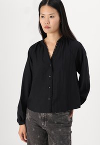 Svart långärmad blus med knäppning fram, gathered halsringning och delikat spetsdetalj, tillsammans med grå jeans med stjärnmönster.