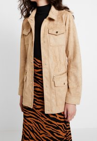 Giacca in faux suede color beige con colletto camicia, due tasche sul petto, due tasche anteriori con zip e dettagli in metallo dorato, abbinata a una gonna a righe arancioni e nere.