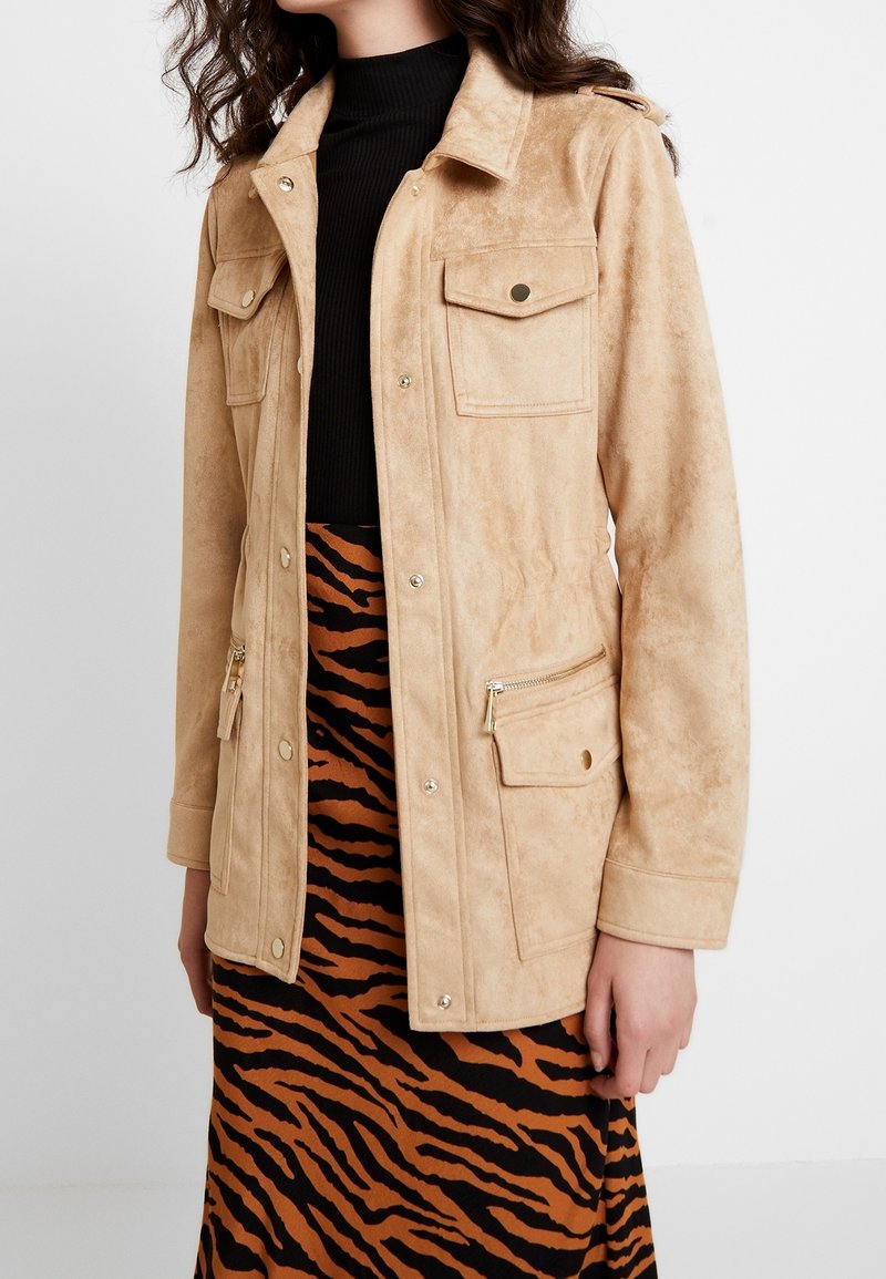 Giacca in faux suede color beige con colletto camicia, due tasche sul petto, due tasche anteriori con zip e dettagli in metallo dorato, abbinata a una gonna a righe arancioni e nere.