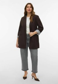 Blazer marron à silhouette allongée, poignets rayés bleu clair, associé à un jean gris raccourci et des mules à talons marron sur un pied.