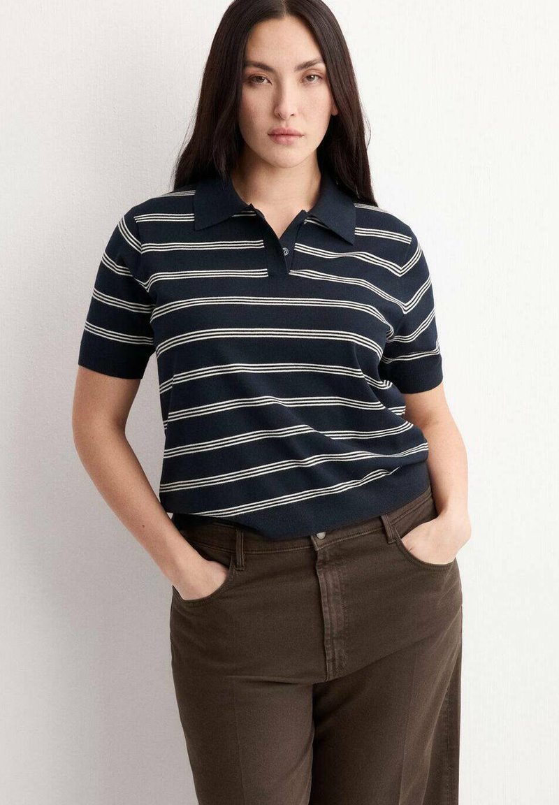 Femme aux longs cheveux foncés portant un polo à manches courtes rayé bleu marine et blanc, un pantalon marron, les mains dans les poches, sur un fond neutre.