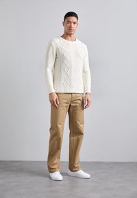 Fülliger cremefarbener Strickpullover mit strukturiertem Rautenmuster, kombiniert mit beigen geraden Hosen und weißen Turnschuhen. Minimaler Hintergrund.