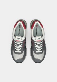 Grå suede- og mesh-sneakers med røde detaljer, hvide snørebånd og en polstret sål. Har "New Balance" logoet fremtrædende på siden.