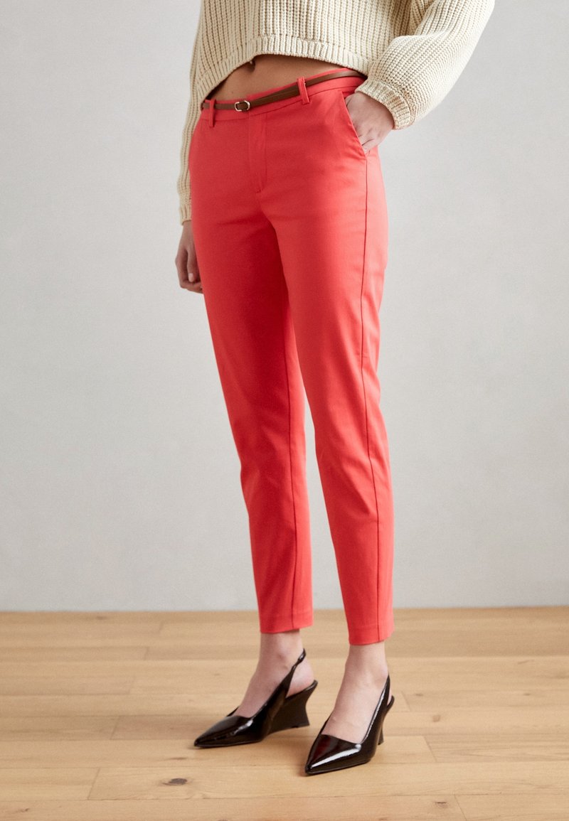Pantalon ajusté couleur corail avec une texture lisse, équipé d'une ceinture élégante et de poches latérales, associé à des talons compensés noirs.