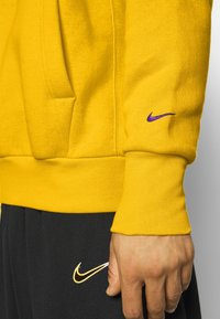 Sweat à capuche jaune Nike avec une poche kangourou, des poignets côtelés et un petit logo violet sur la manche, associé à un pantalon noir avec un logo.