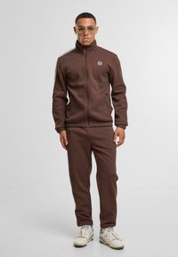 Brūna sporta uzvalka ar rāvējslēdzēju, izgatavots no mīksta auduma, ar augstu apkakli, sānu svītrām un pieguļošu dizainu. Apvienots ar baltām sporta kurpēm.