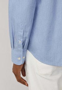 Lichtblauwe langslijf shirt met een gestructureerde stof, met knoopmanchetten en een gebogen zoom. Gecombineerd met witte broek, met details van zijzakken.