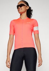 Lys rosa sykkeltrøye med kort glidelås, med en hvit aksentstripe på ermet. Sorte polstrede shorts kompletterer antrekket.