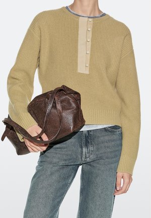 Persona che indossa un maglione a coste beige e jeans grigi, che tiene una grande borsa in pelle marrone scuro con trama.