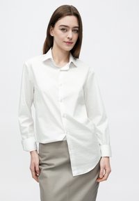 Νεαρή γυναίκα με ίσια καστανά μαλλιά φοράει λευκό πουκάμισο με κουμπιά και μπεζ φούστα, στέκεται μπροστά σε απλό λευκό φόντο.