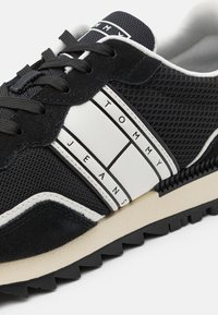 Zapatilla negra con materiales de malla y gamuza, que presenta rayas de marca blancas, una suela textura negra y una forma atlética clásica.