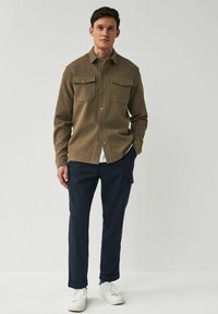 Camisa verde oliva de botones con dos bolsillos en el pecho, combinada con pantalones cargo azul marino y zapatillas blancas. Diseño simple y moderno con textura de tela.