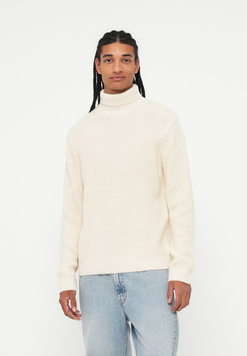 Maglione a collo alto intrecciato di colore crema con una vestibilità ampia. Polsini e orlo a coste, con un motivo testurizzato. Abbinato a jeans in denim blu chiaro.