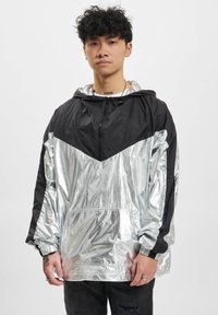 Silberne und schwarze Kapuzenwindjacke mit einem eleganten, glänzenden Finish, ausgestattet mit einem Reißverschluss vorne, elastischen Bündchen und einem diagonalen Farbblockdesign.