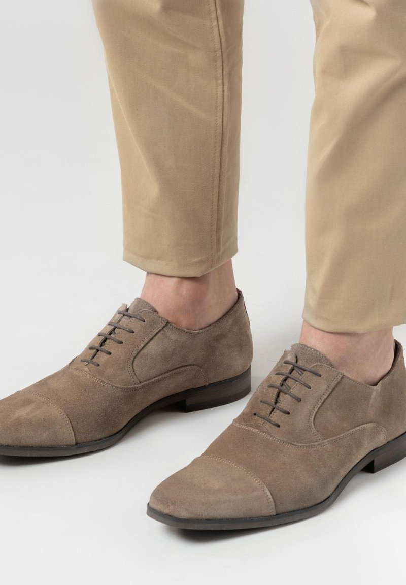 Suède bruine jurk schoenen met een cap toe ontwerp, vetersluiting en een lage houten hak, gecombineerd met beige broeken.