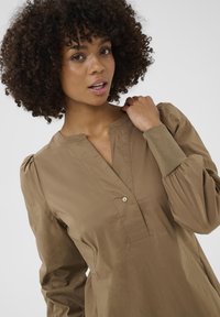 Göndör hajú nő, aki barna, hosszú ujjú blúzt visel, gombos V-nyakkal, egyik kezével a gallérját fogja egy egyszínű háttér előtt.