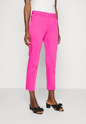 Kobieta w jasnoróżowych spodniach o kroju typu cropped z bocznymi kieszeniami oraz czarnych sandałach na obcasie z kokardą, na jednolitym białym tle.