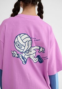 Lyserød oversized t-shirt med korte ærmer, der har et grafisk motiv af en tegneseriefigur. Blå detaljer på de nederste ærmer og en blød tekstur.