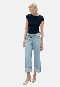 Navy fitted top, high-waisted light blue wide-leg jeans with a decorative belt. White open-toe heels accent the outfit.