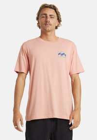 Jasnoróżowa bawełniana koszulka z krótkim rękawem, okrągłym dekoltem i małą niebieską łatą w wzory fal na lewej piersi.