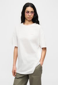 Γυναίκα με μακριά σκούρα μαλλιά που φοράει ροζ γυαλιά ηλίου, μια άνετη λευκή μπλούζα Columbia και χακί παντελόνια, με το ένα χέρι στην τσέπη.