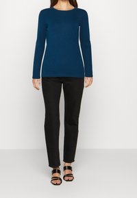 Top de manga larga en color navy, hecho de una tela suave y elástica, combinado con jeans negros ajustados y sandalias negras de tiras sobre un fondo neutral.