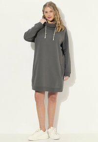 Graues Sweatkleid mit Kapuze, Kordelzug-Details, langen Ärmeln und Seitentaschen. Kombiniert mit weißen Sneakers. Glatte Textur.