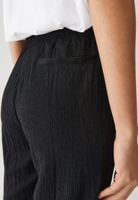 Zwarte textuur broek met een geplooid ontwerp, voorzien van een achterzak en een zachte, elastische tailleband. Close-up van de zijkant.