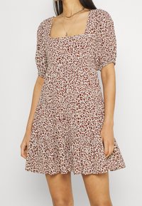 Leopardi-kuosiset mekko pehmeässä kankaassa, jossa on neliömainen kaula-aukko, puffatut lyhyet hihat ja kerroksellinen hame. Maalauksellinen väriteema ruskeilla vivahteilla.