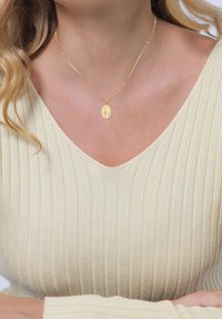 Goldene Halskette mit einem ovalen Anhänger, der ein strukturiertes Design aufweist, kombiniert mit einem ribbeligen, hellbeigen Oberteil. Schlichte, elegante Ästhetik.