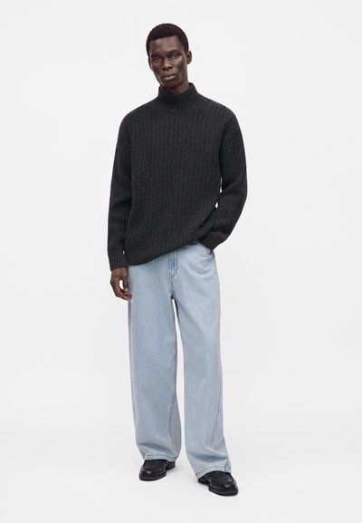 Homme portant un pull en maille à col roulé gris foncé, un jean bleu clair à jambes larges et des chaussures noires, debout devant un fond blanc uni.